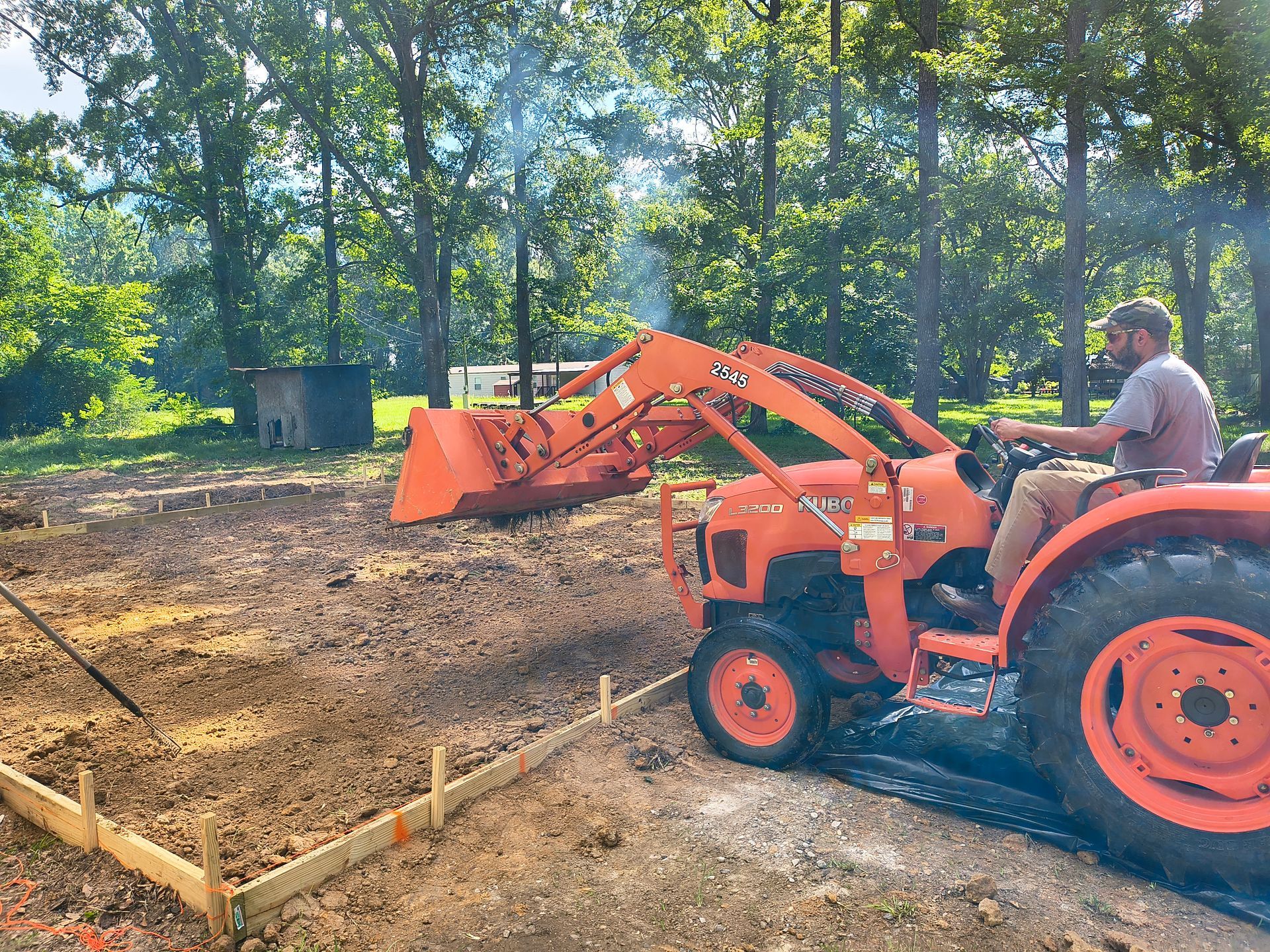 Man operating orange tractor leveling soil in a yard, with a wooded background.
