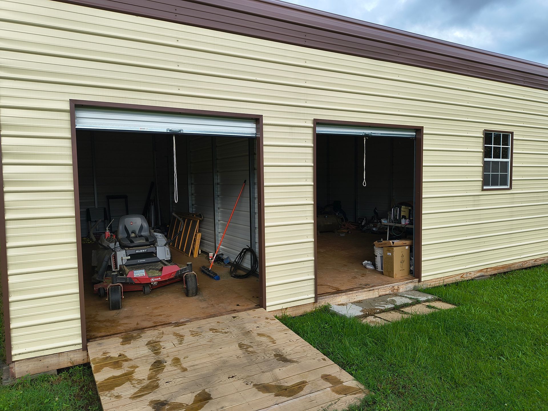 Two garage doors open, showing interior with lawnmower and tools; building has beige siding and brown roof.