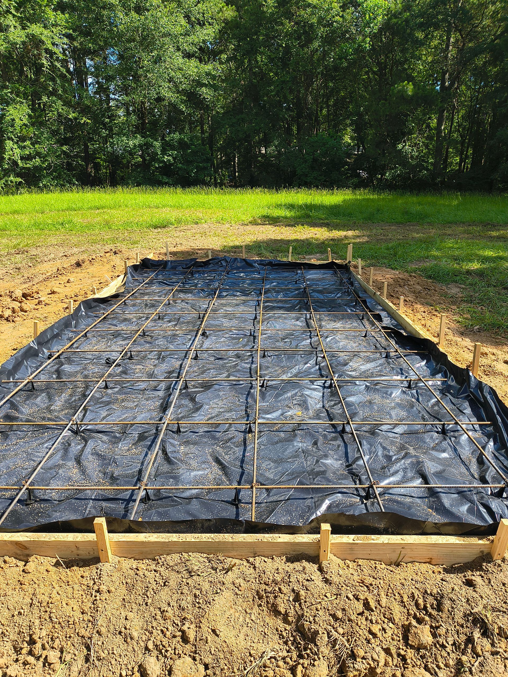 Construction site: wooden frame, rebar grid on black fabric, surrounded by dirt, in front of green grass and trees.