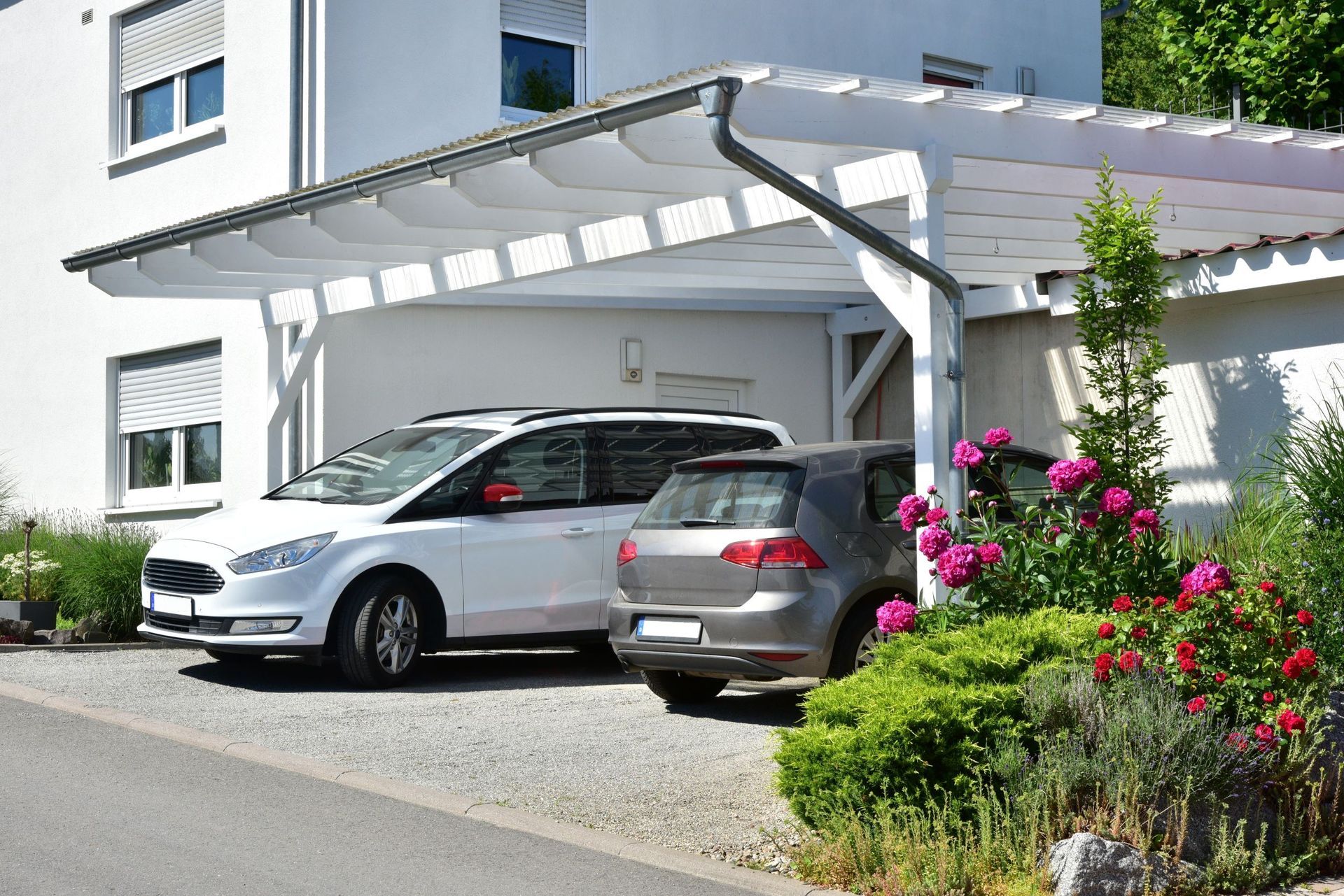 Cars parked under a white carport attached to a house, gravel driveway, with greenery.