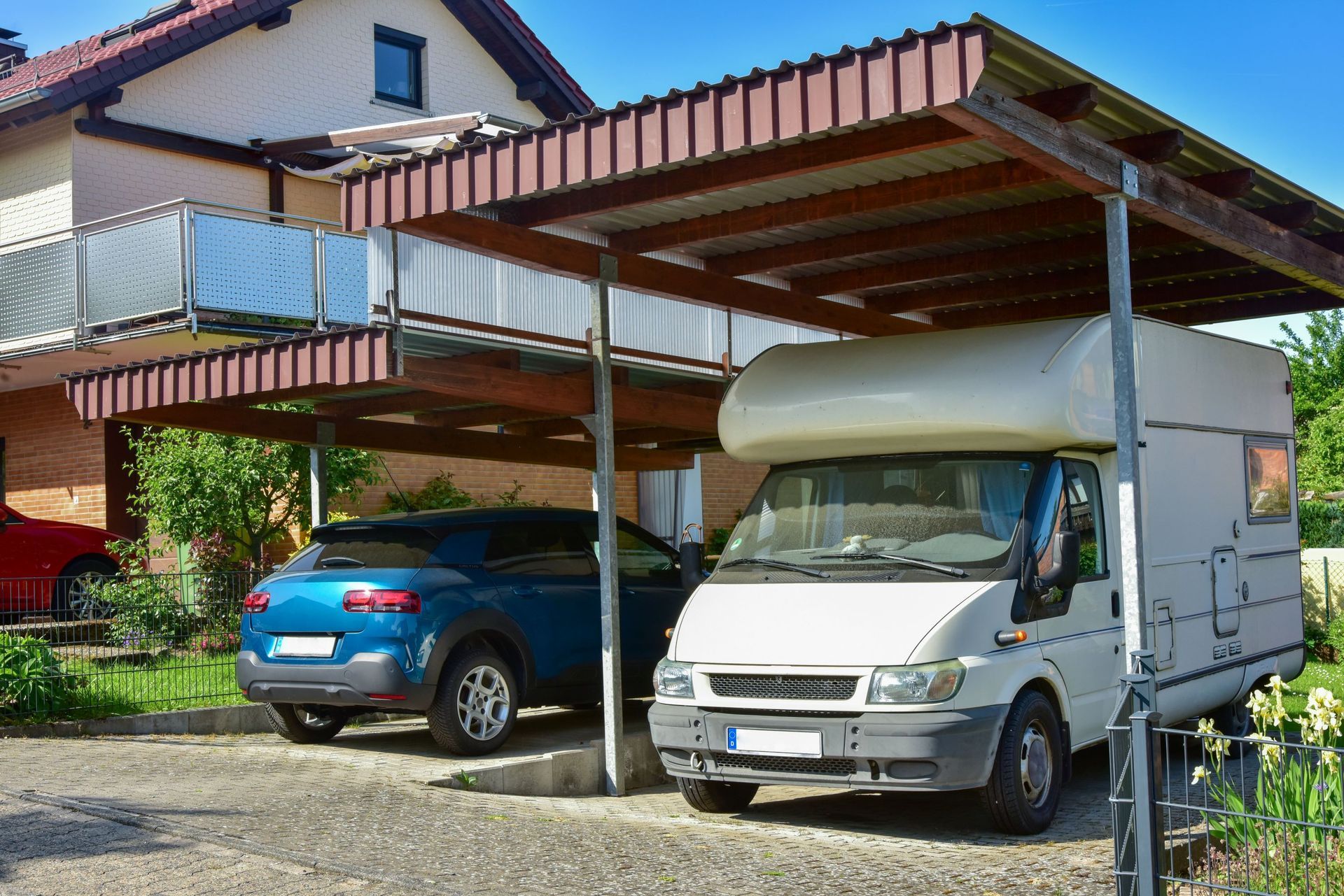 A car and a campervan parked under a brown corrugated metal carport next to a house.