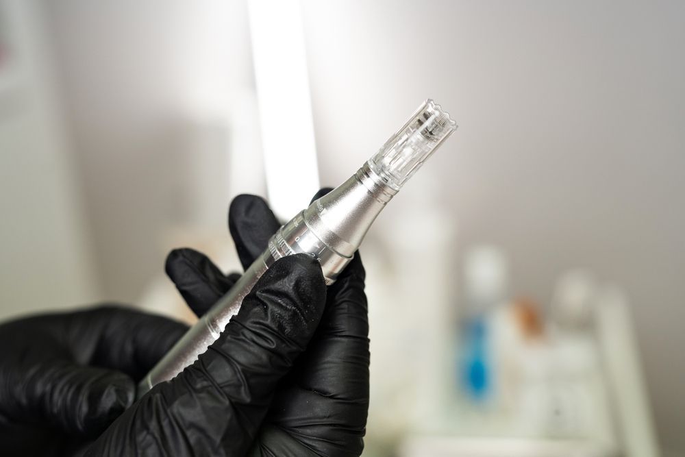Hand holding a silver permanent makeup pen with a needle. Black gloved hands, white background.