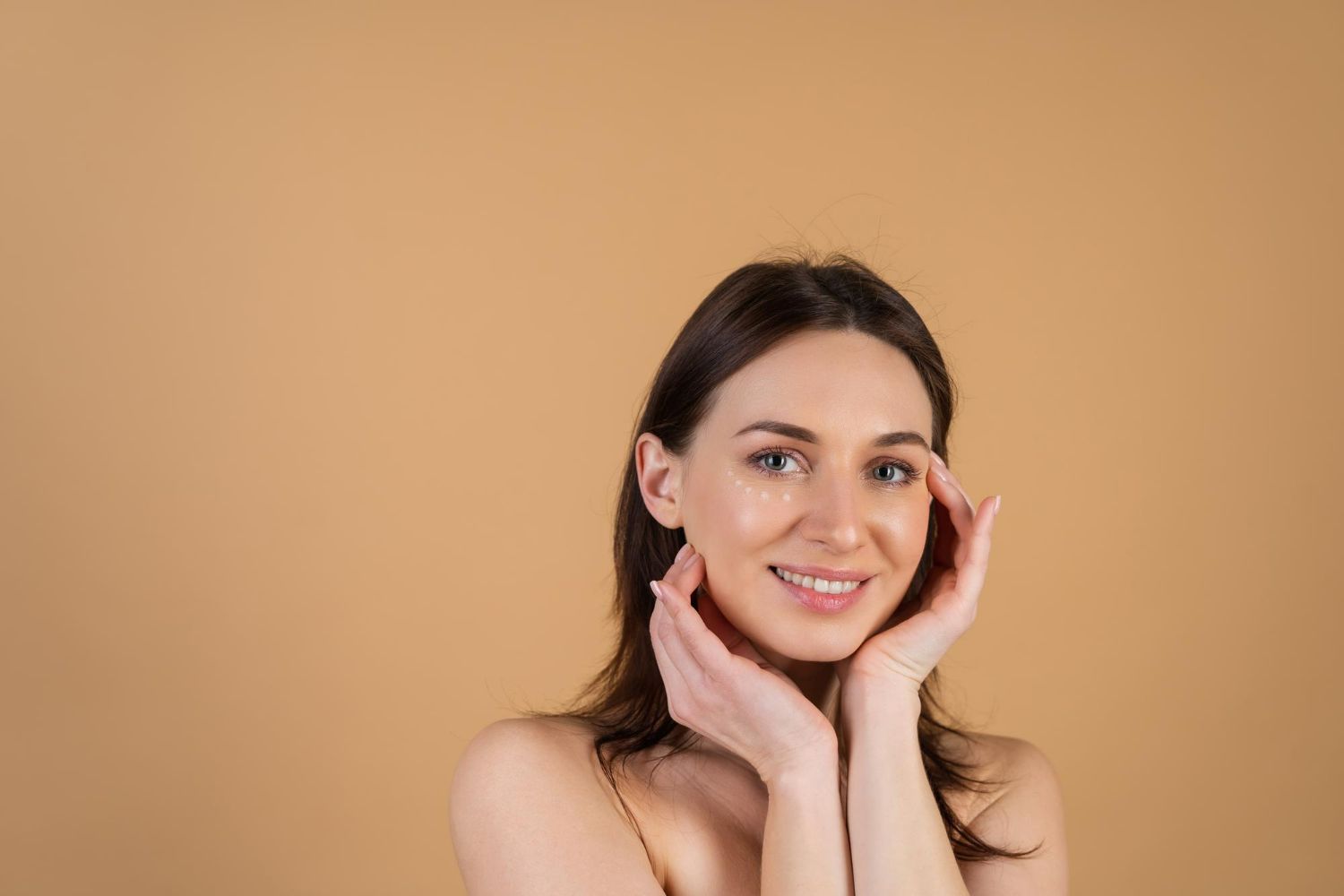 Smiling woman with light skin, dark hair, hands near face, tan background.