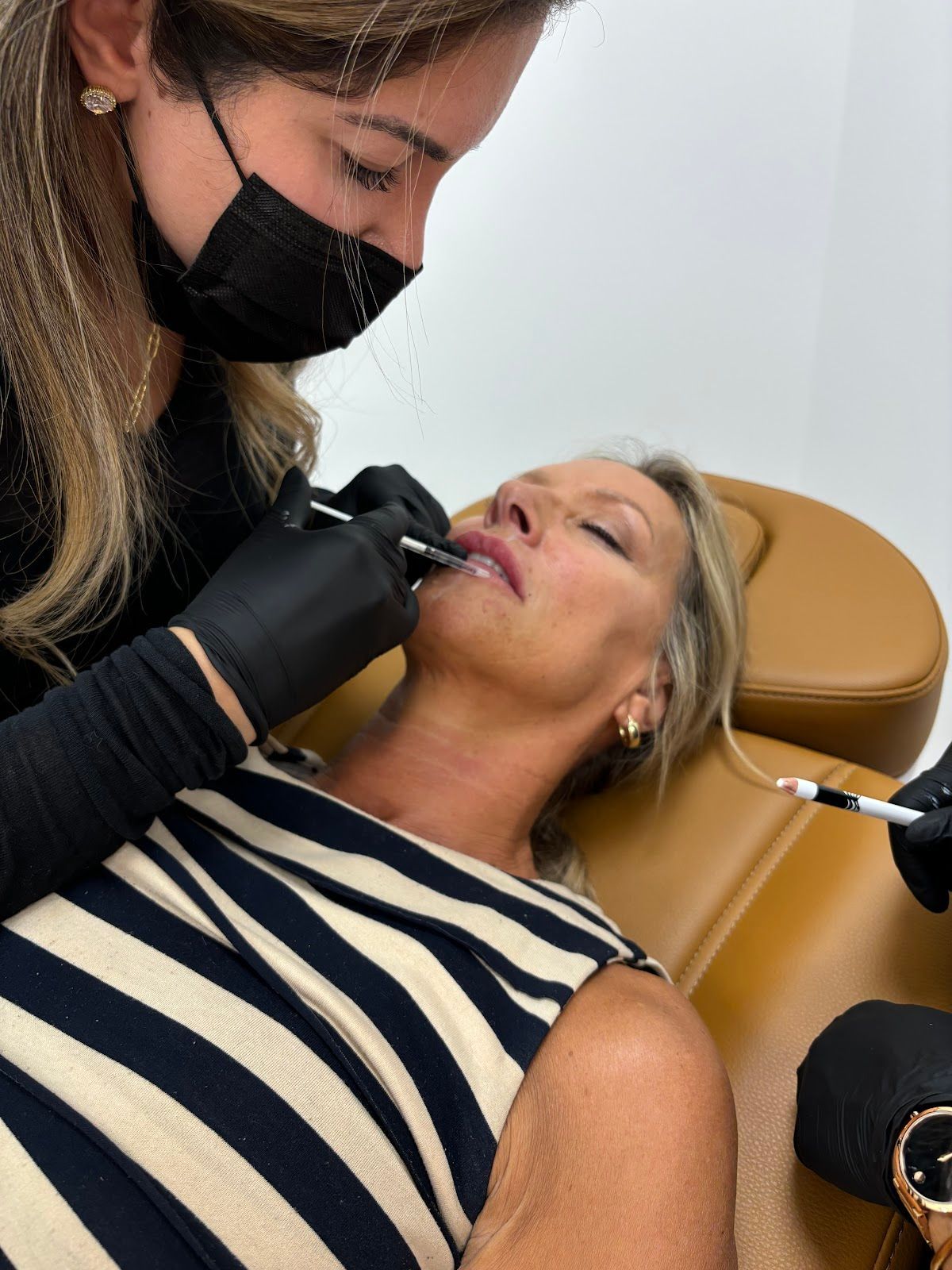 A woman receives lip filler injections from a masked practitioner. The patient is in a medical office setting.