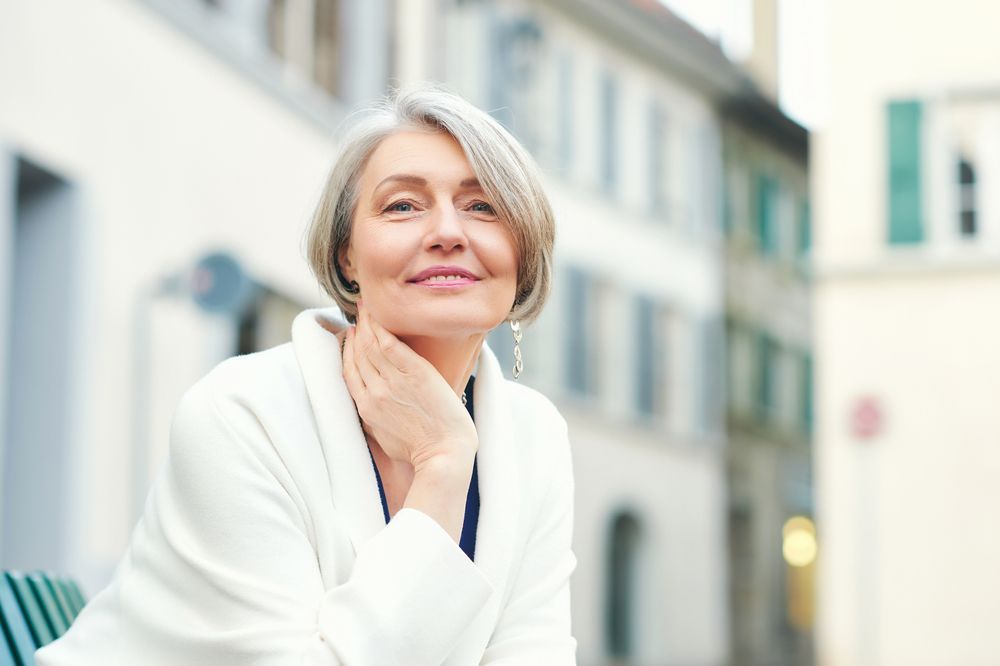 A woman with gray hair is sitting on a bench in front of a building.