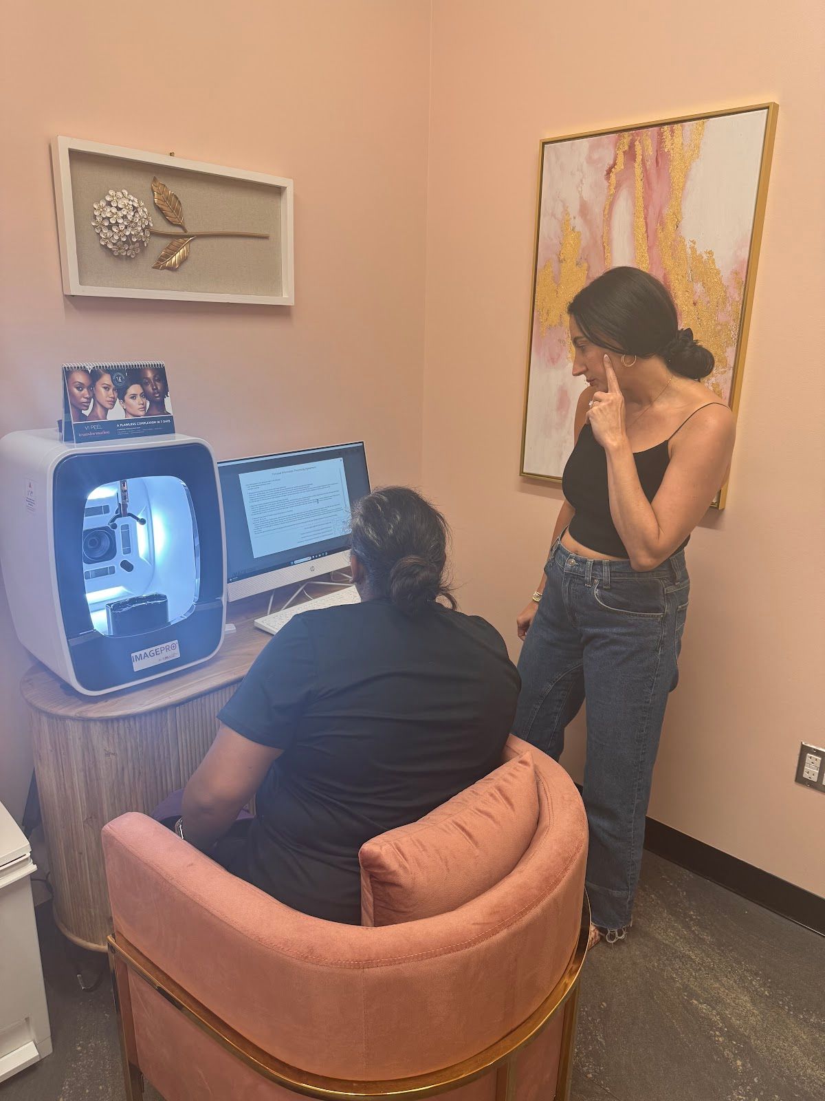 Two women in a pink-walled office, one operating a dental machine, the other observing.
