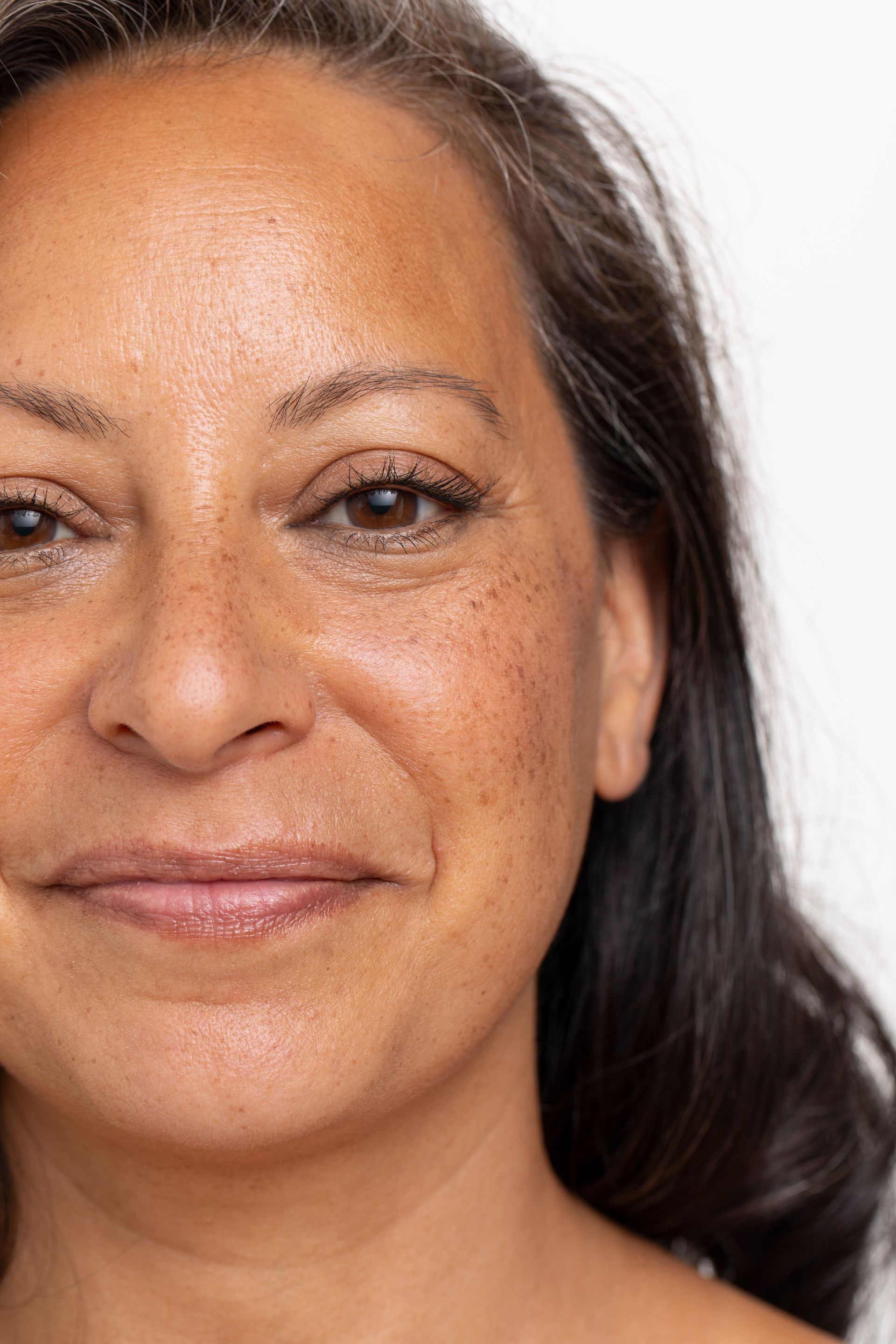 Woman with brown skin, dark hair, and freckles smiles at the camera against a white backdrop.