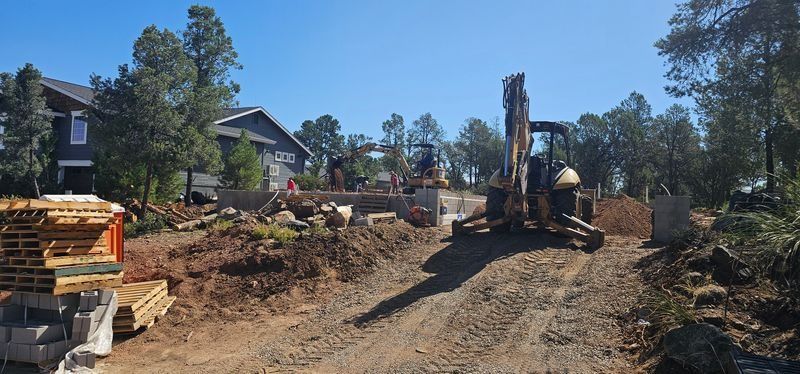 Construction site with excavator clearing dirt beside stacked lumber and houses under a blue sky