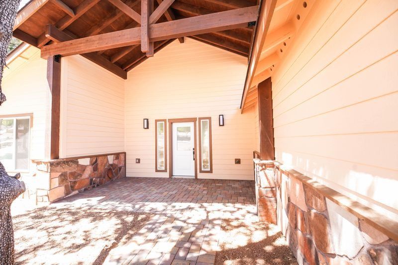 Covered porch with wooden beams, white siding, stone base, and double glass doors in bright sunlight