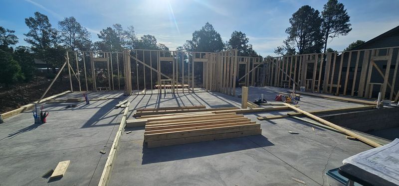Partially framed house foundation with lumber stacks and construction materials under a bright blue sky