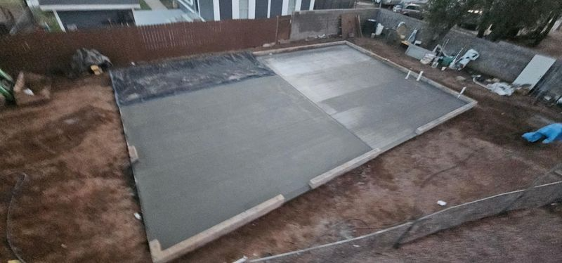 Backyard construction site with a rectangular concrete slab and wooden formwork, surrounded by dirt and fencing
