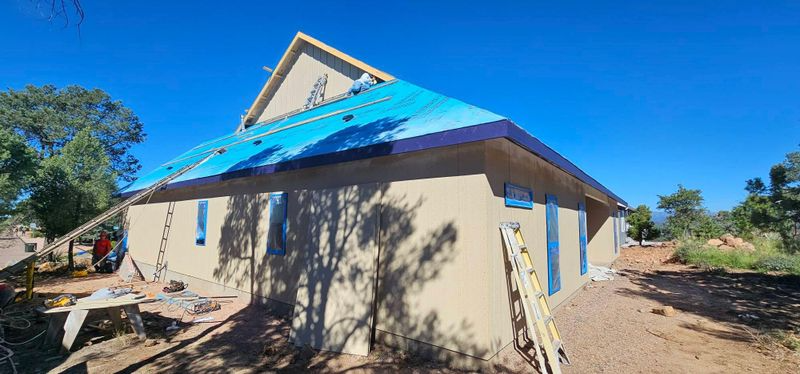 Partially built house with blue roof, beige walls, and ladder in a sunny dirt yard