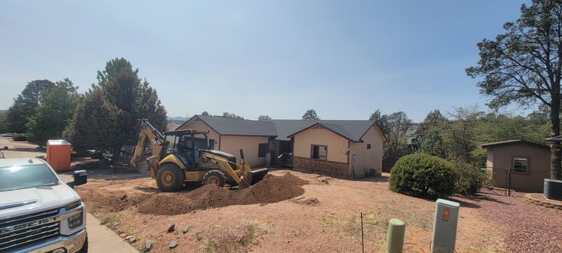Backhoe and truck in a dusty yard beside a single-story house and trees under a clear sky