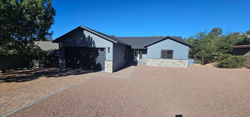 Single-story gray house with a garage and gravel driveway in a sunny suburban setting
