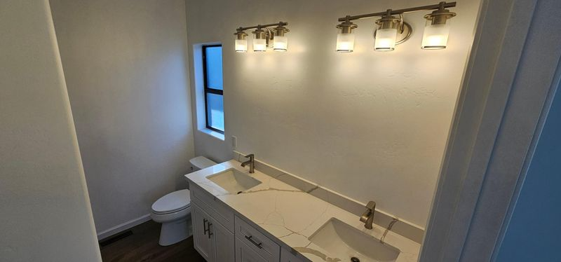 Modern bathroom vanity with double sinks, wall lights, mirror, and a toilet by a small window