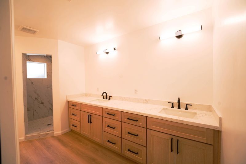 Empty bathroom vanity with double sinks, wooden cabinets, and wall sconces lit above a shower door.