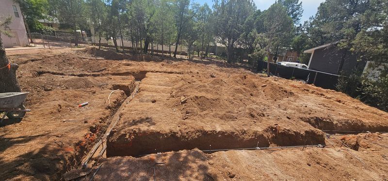 Sunlit dirt lot with freshly leveled soil, trees in the background, and a small building on the right.