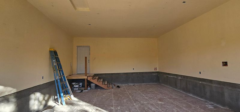 Empty beige room under renovation with ladders, scattered tools, and dark baseboard trim