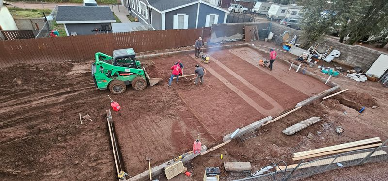 Workers paving a red-brown driveway as a green tractor levels soil on a residential lot.