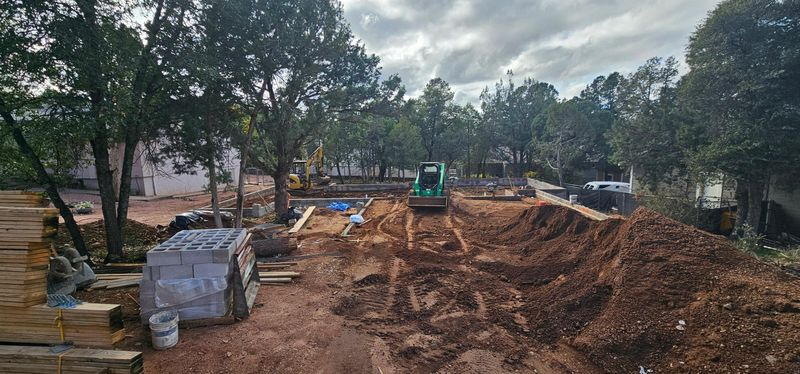 Construction site with excavator digging a large dirt trench beside trees and stacked materials