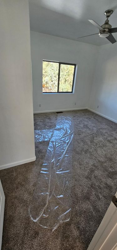 Empty small bedroom with gray carpet, white walls, ceiling fan, and a window letting in daylight.