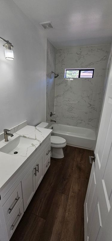 Small white bathroom with vanity, toilet, bathtub, and frosted window on dark wood floor