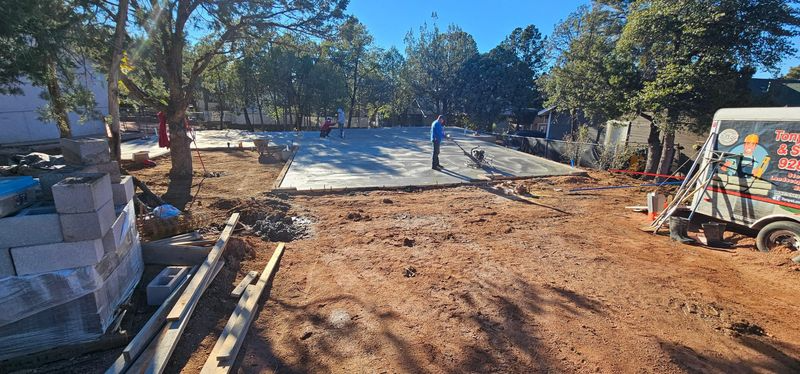 Construction site with dirt lot, workers, and equipment under trees and blue sky