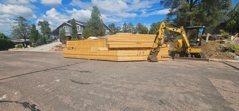 Yellow lumber pile beside a yellow excavator on a roadside under a blue sky