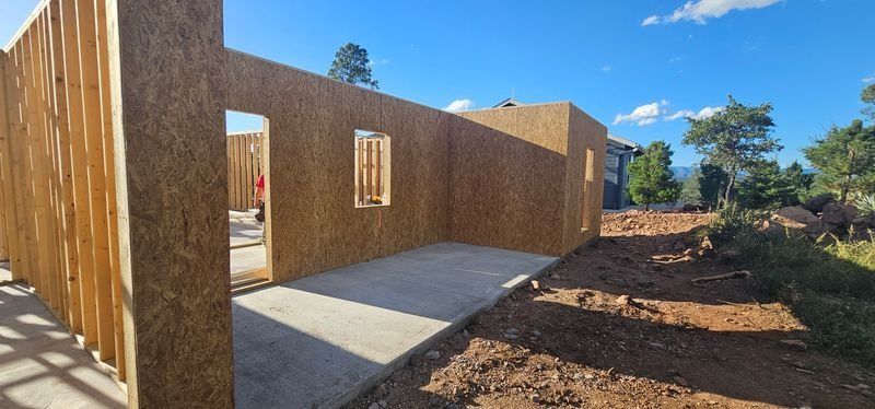 Partially built wooden house framing with concrete slab and dirt yard under blue sky