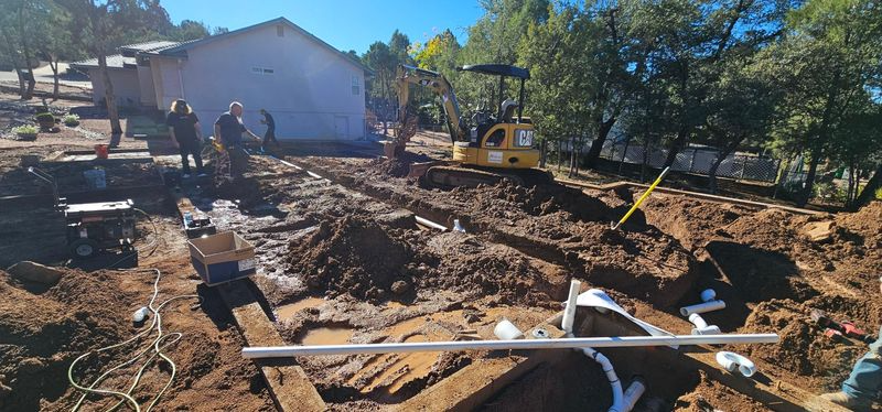 Backyard construction site with a large dirt excavation, tools, and workers near a house under sunny skies