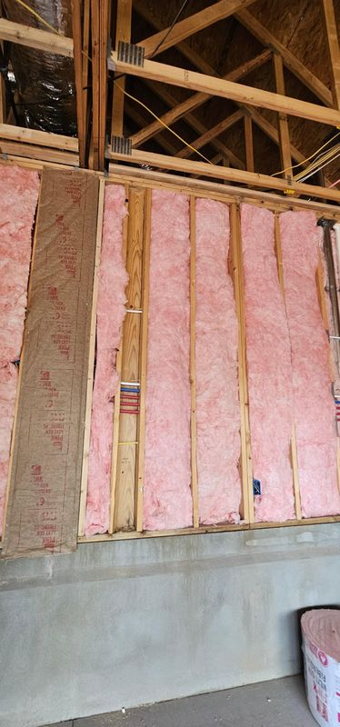 Interior wall under construction with pink insulation batts between exposed wooden studs and roof trusses.