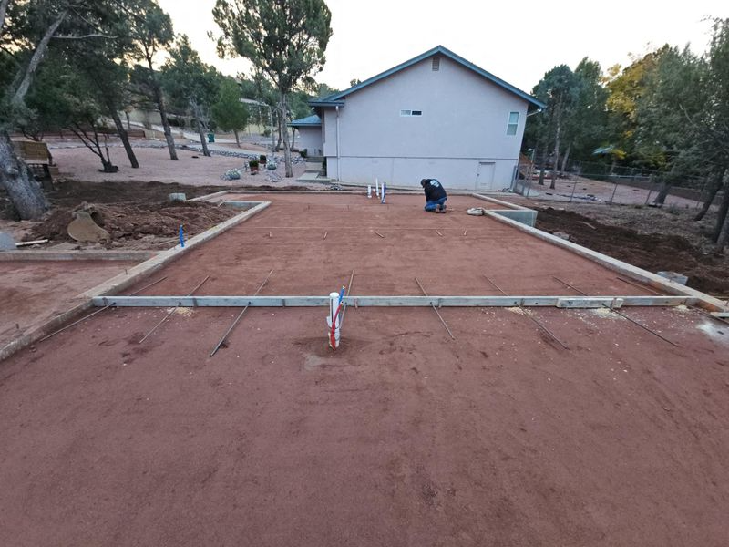 Roughly graded dirt area with wooden frame and white house in the background, surrounded by trees.