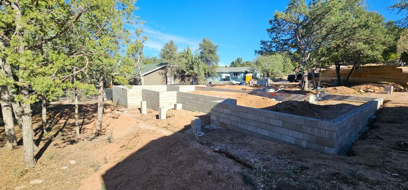 Construction site with concrete foundation walls and dirt lot under trees, with an overpass in the background