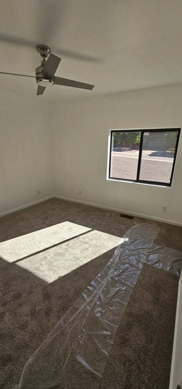 Empty unfurnished room with ceiling fan, window, and sunlight on carpeted floor.