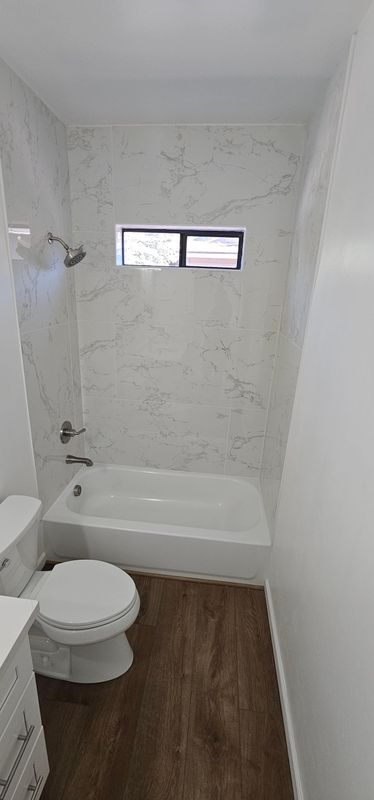 Small bathroom with shower-tub, toilet, wood-look floor, and a small window above the tub.