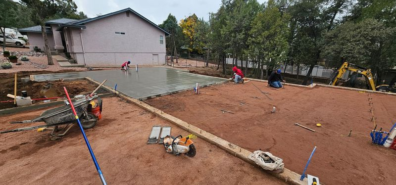 Construction site with a house foundation, wooden forms, pipes, and an excavator in the background
