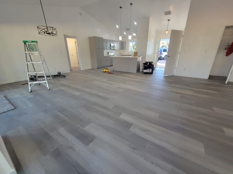 Empty room with gray wood flooring, white walls, ladder, and kitchen area in back