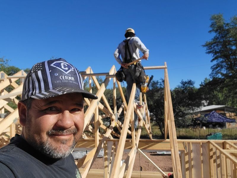 Man smiling in foreground at a construction site, with a worker standing on a wooden frame behind him under a blue sky.