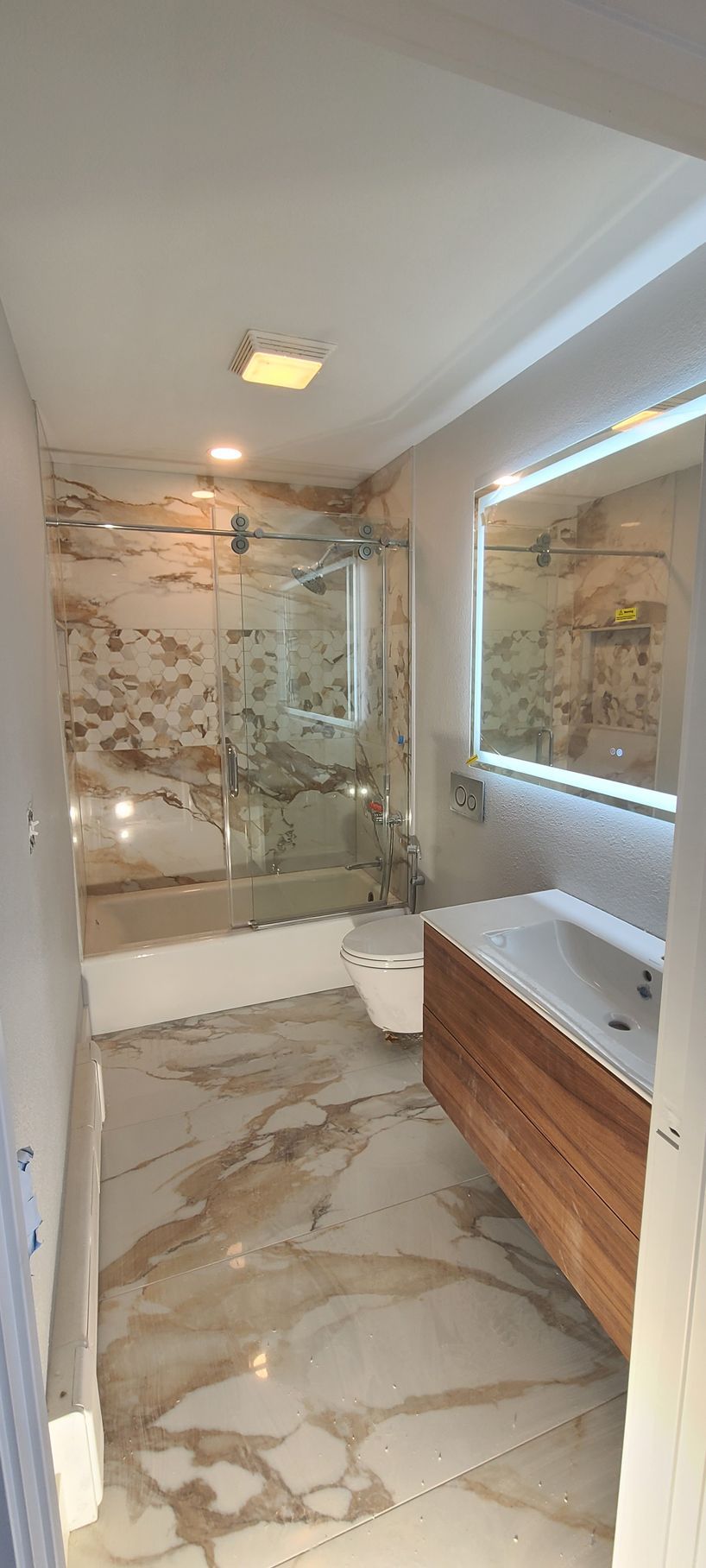 Modern bathroom with marble-patterned tile and walls. Features a floating vanity, glass shower door, and lit mirror.