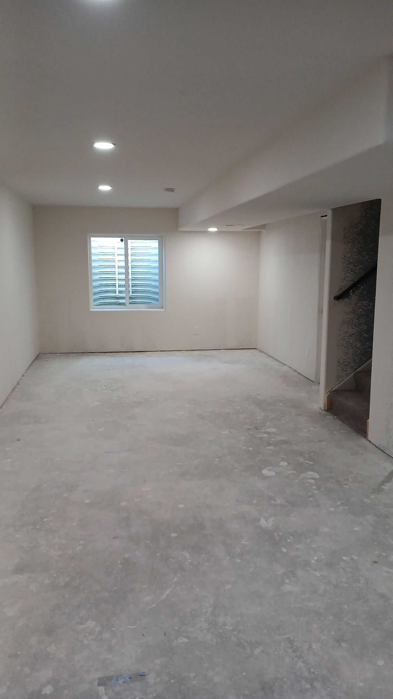 Empty room with unfinished concrete floor, window, and recessed lights. Unfinished doorway on right.