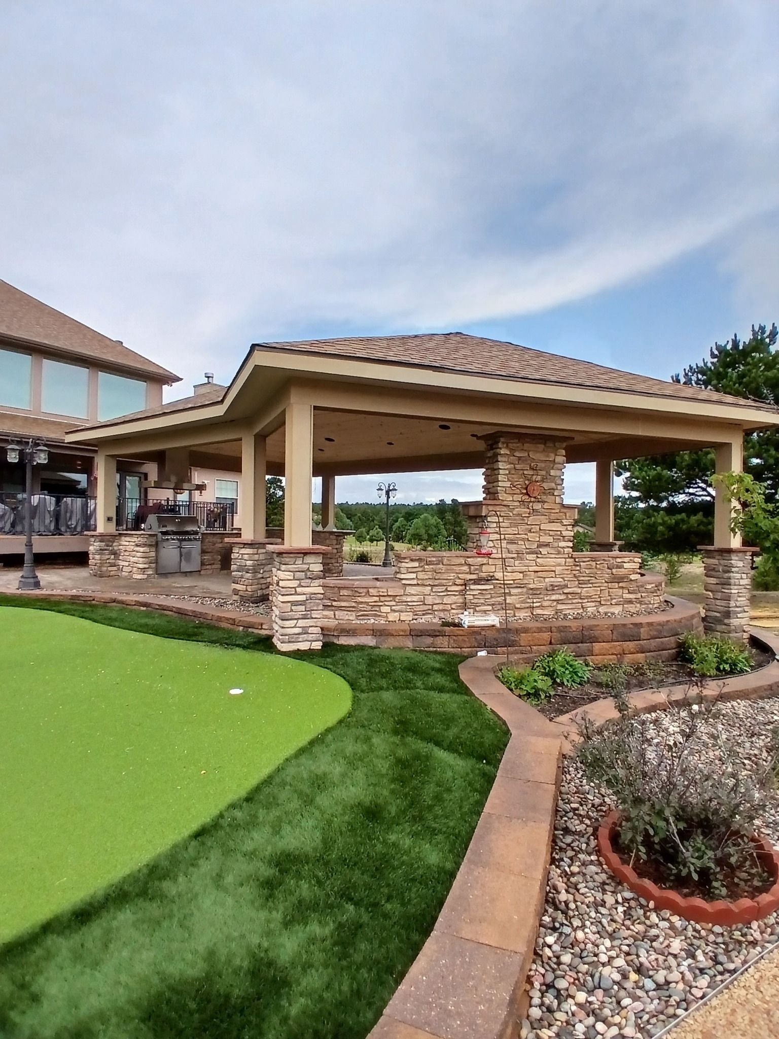 Gazebo with stone accents and tile roof next to a putting green, golf course in background.