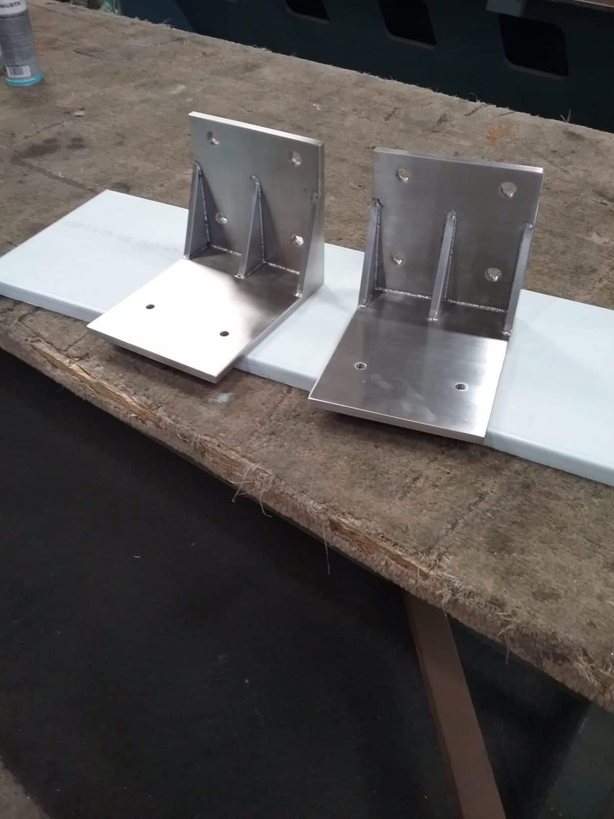 Two Shiny Metal Brackets on a White Surface, Resting on a Brown Table — Highlander Metals in Nowra, NSW