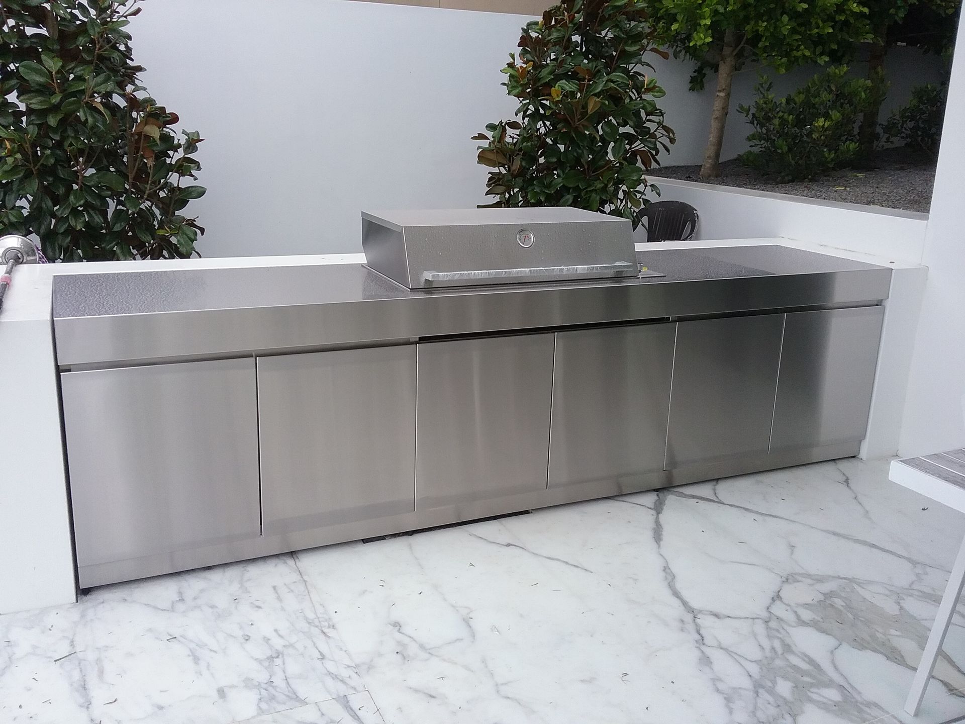 A stainless steel outdoor kitchen island with a built-in grill on a marble patio against a white wall with greenery — Highlander Metals in Kirrawee, NSW