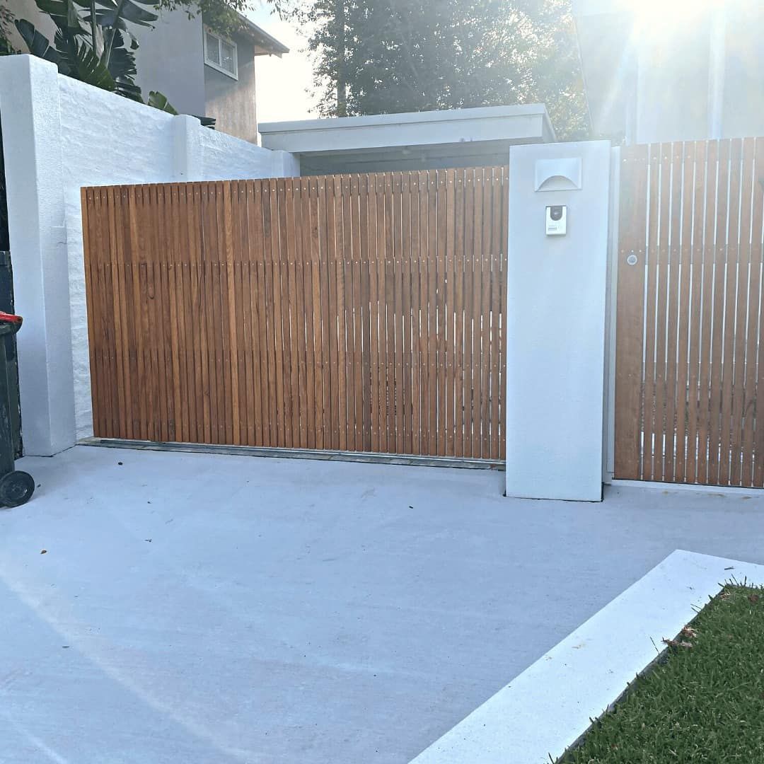 A modern driveway gate made of vertical wooden slats, set between two white stucco pillars with an intercom system — Highlander Metals in Kirrawee, NSW