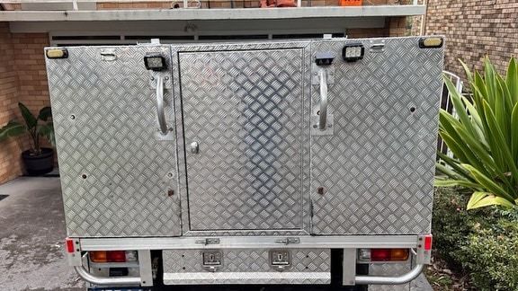 A diamond-plate aluminum truck canopy with a central door, mounted handles, and rear lights, parked outdoors — Highlander Metals in Kirrawee, NSW