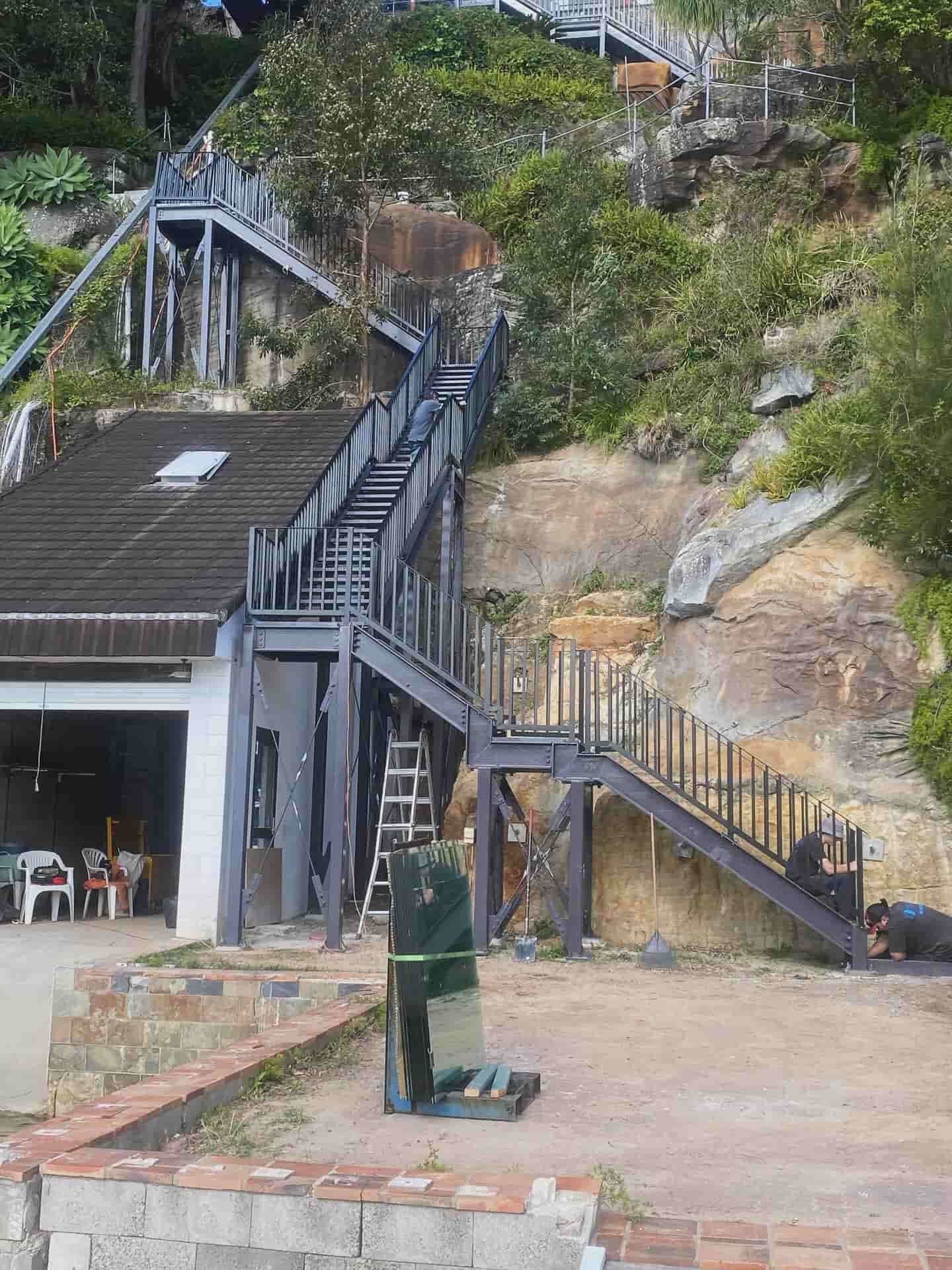 A multi-level metal outdoor staircase ascends a steep, rocky hillside next to a house with a dark tiled roof — Highlander Metals in Kirrawee, NSW