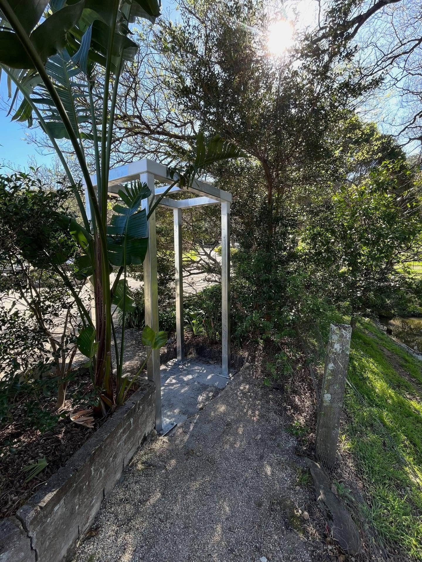 An outdoor metal shower frame stands on a concrete pad surrounded by trees and greenery in a sunny, wooded area — Highlander Metals in Kirrawee, NSW