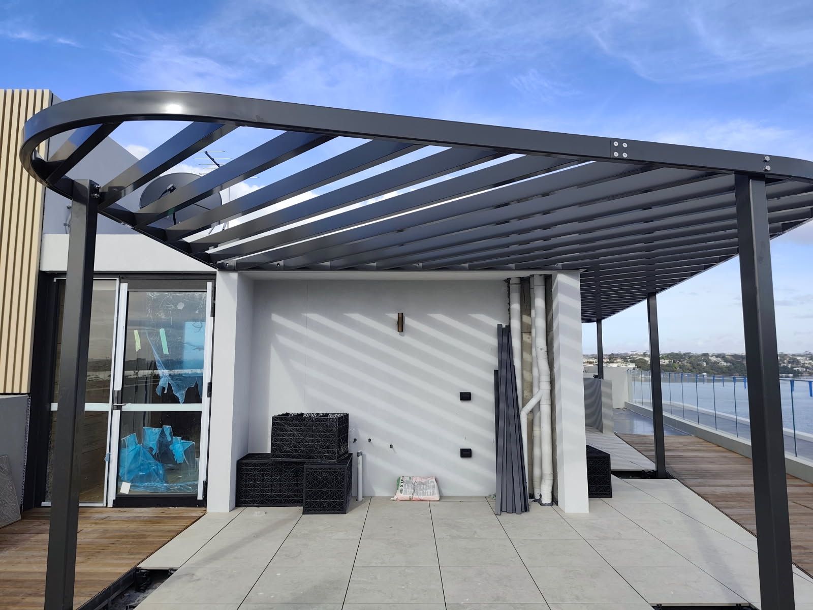 A modern dark-framed patio pergola with slatted overhead shading on a tiled rooftop deck overlooking a body of water — Highlander Metals in Kirrawee, NSW