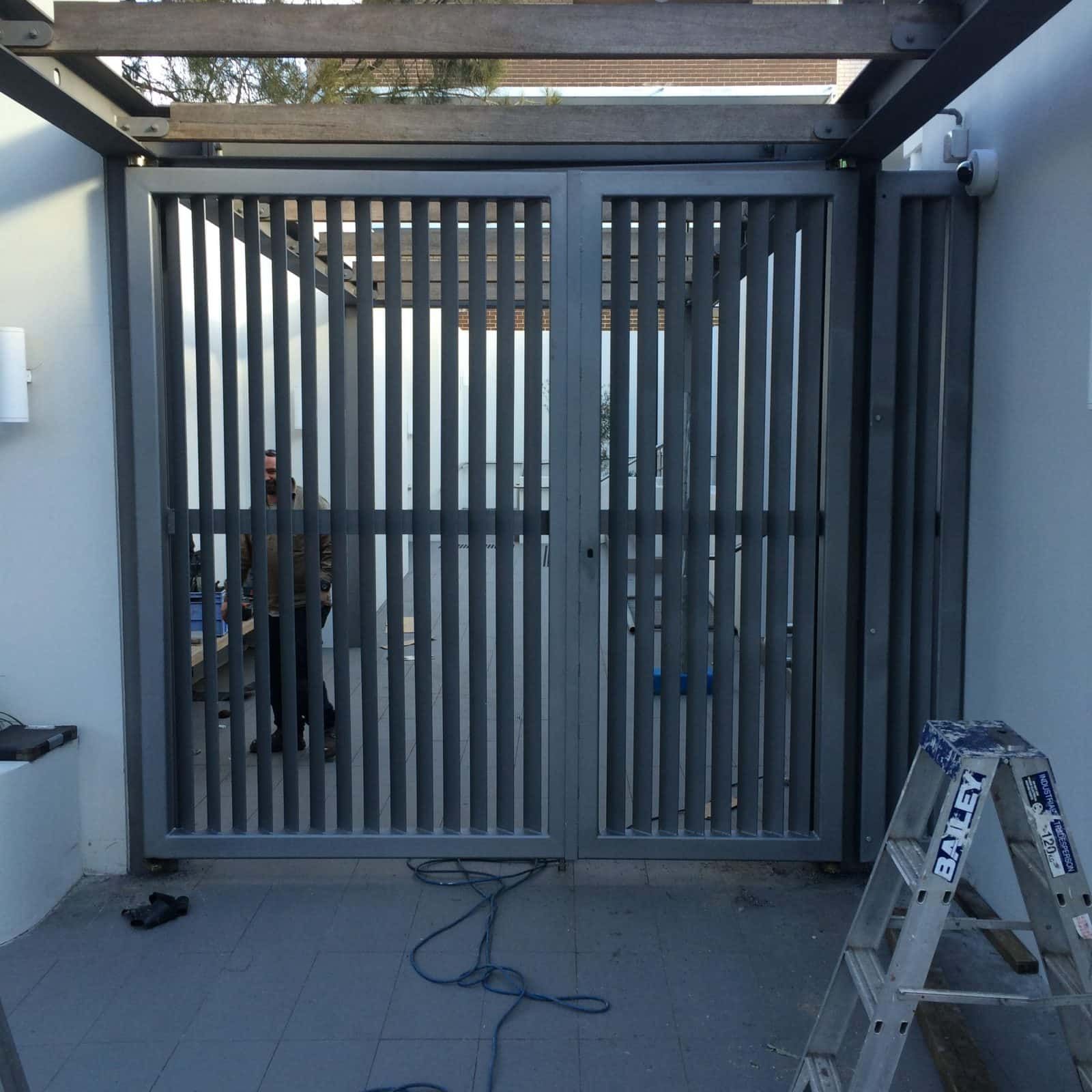 A gray, slatted metal security gate installed between two white walls, with a ladder and power cords on the ground — Highlander Metals in Kirrawee, NSW