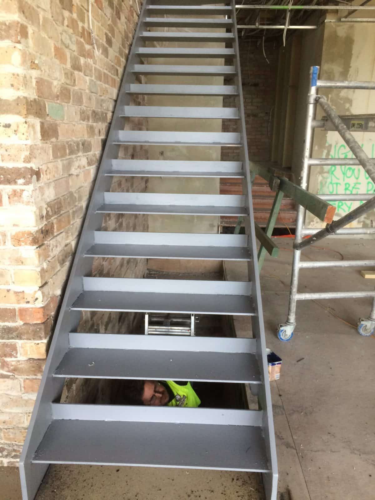 A person wearing a high-visibility vest works in a crawl space beneath a set of newly painted gray stairs — Highlander Metals in Kirrawee, NSW
